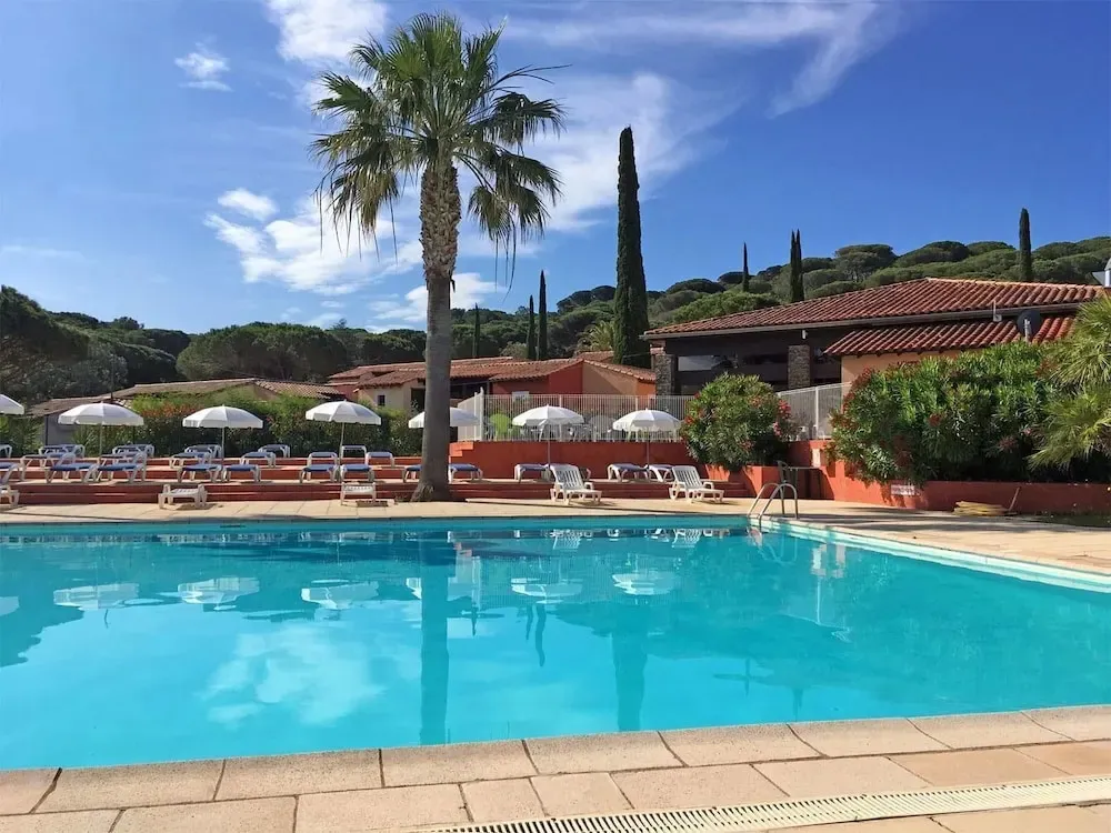 Une piscine turquoise reflète un bâtiment aux toits de tuiles rouges et des palmiers sous un ciel bleu.