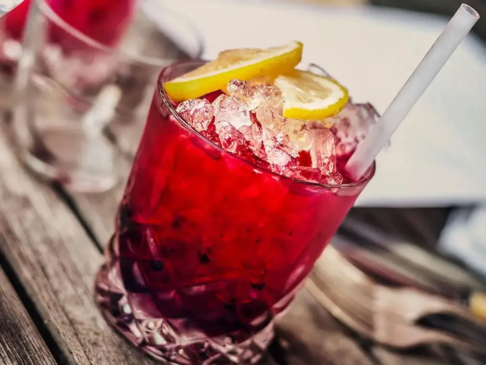 Un cocktail rouge glacé est servi dans un verre décoré, garni d'une tranche de citron et d'une paille.
