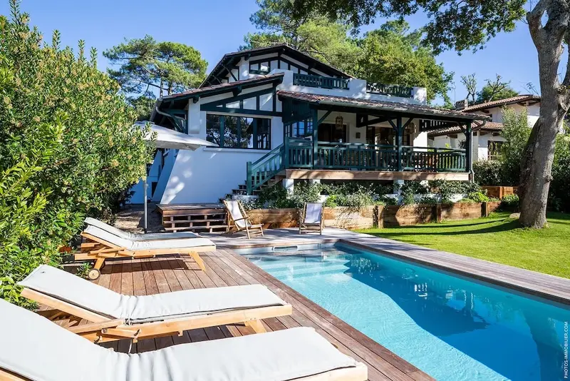 Une piscine bordée de chaises longues devant une maison blanche à colombages.