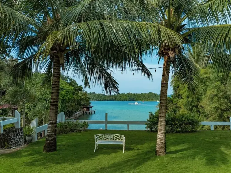 Un banc blanc se trouve sur une pelouse verte encadrée par des palmiers, avec vue sur une baie turquoise.