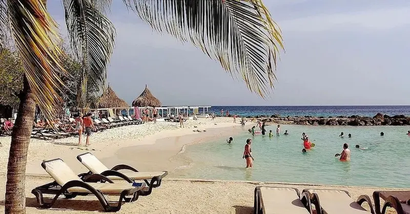 Une plage de sable blanc bordée de transats et de palmiers, avec des baigneurs dans une eau turquoise.