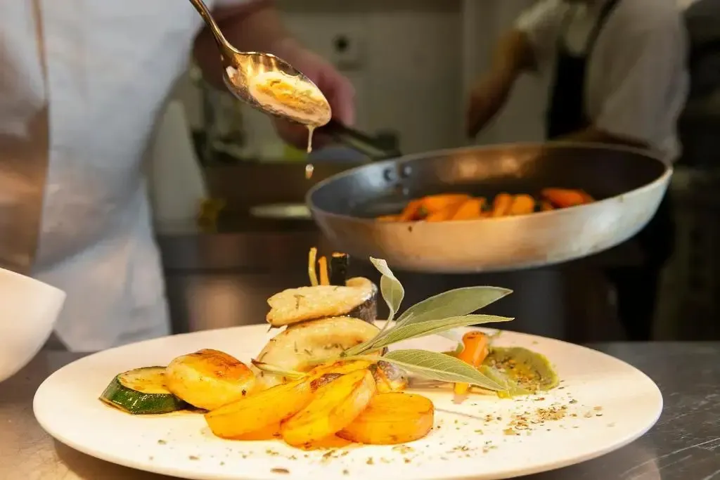 Un plat de légumes et de viande est dressé sur une assiette blanche, tandis qu'une sauce est versée depuis une louche.