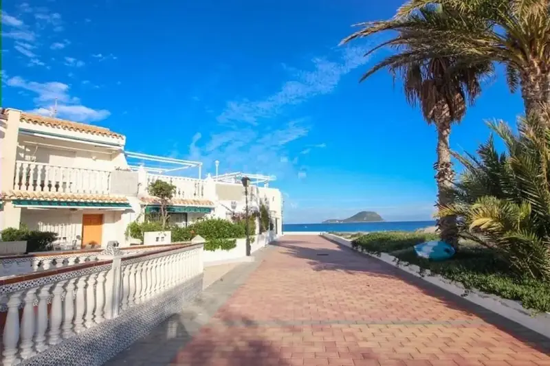 Allée piétonne bordée de bâtiments blancs et de végétation luxuriante, avec vue sur la mer et les montagnes au loin.