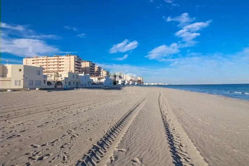 Une longue plage de sable fin s'étend devant des bâtiments côtiers sous un ciel bleu nuageux.