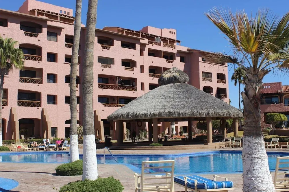 Une piscine turquoise bordée de transats devant un bâtiment rose à balcons.