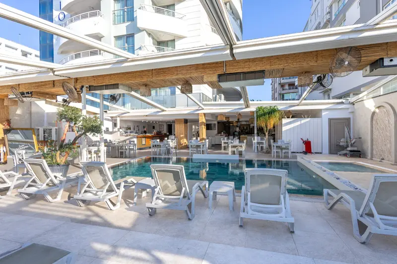 Transats blancs disposés autour d'une piscine rectangulaire sous une pergola en bois.