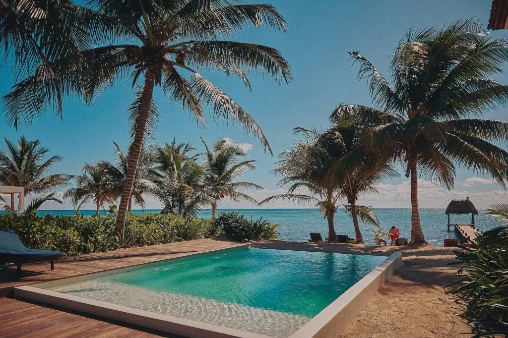 Une piscine turquoise bordée d'une terrasse en bois, avec des palmiers et une vue sur la mer.