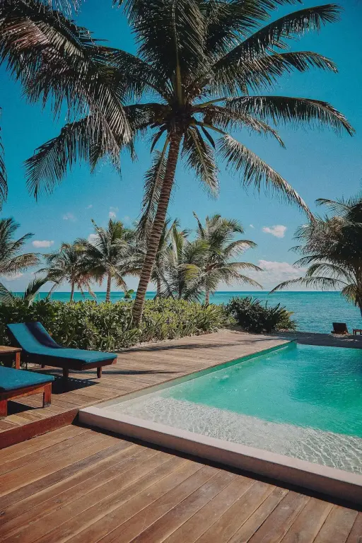 Transats bleus disposés au bord d'une piscine turquoise, palmiers et mer turquoise en arrière-plan.