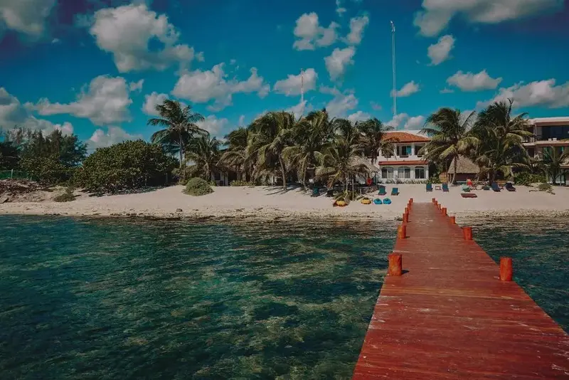 Un ponton en bois mène vers une plage bordée de végétation et de bâtiments.