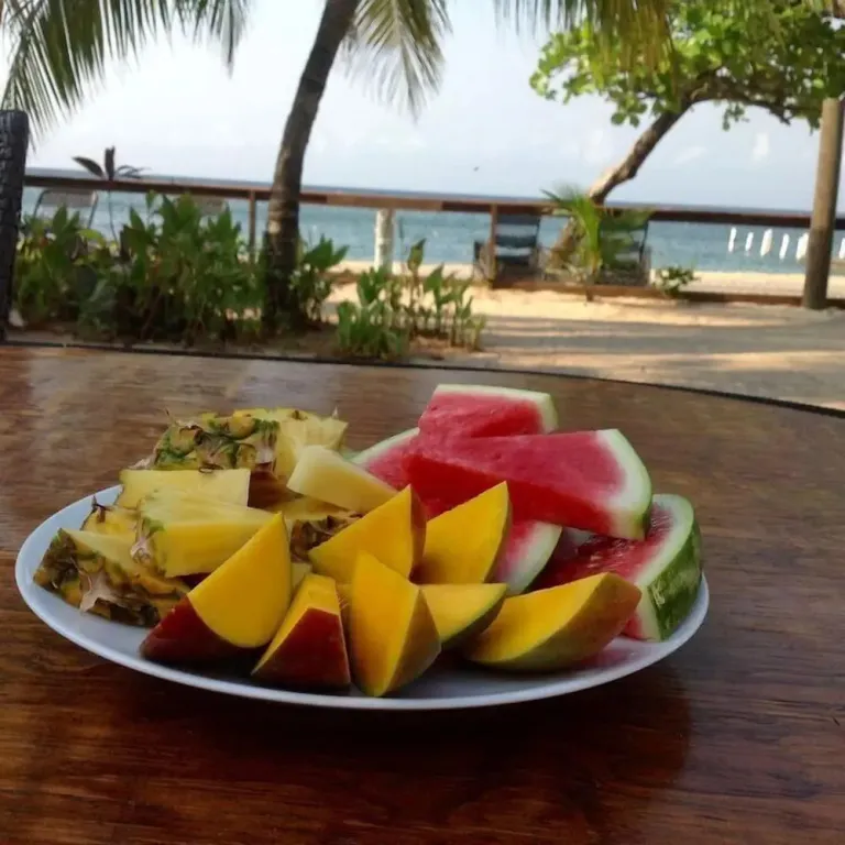 Une assiette de fruits frais est présentée sur une table en bois avec vue sur une plage tropicale.