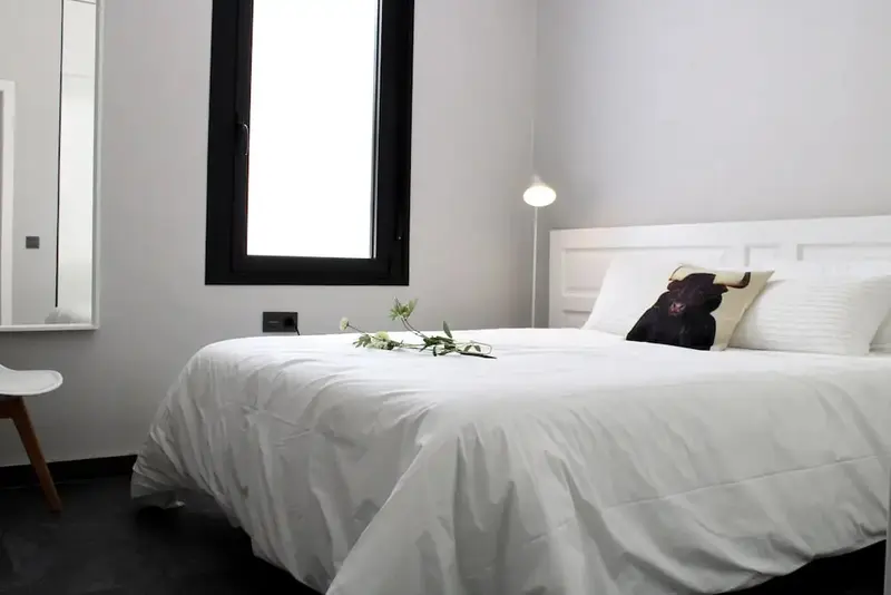 Une chambre lumineuse avec un lit recouvert de linge blanc et une fenêtre encadrée de noir.