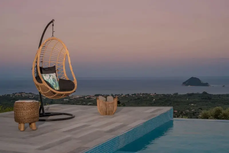 Un fauteuil suspendu en osier se trouve au bord d'une piscine avec vue sur la mer et un paysage montagneux.