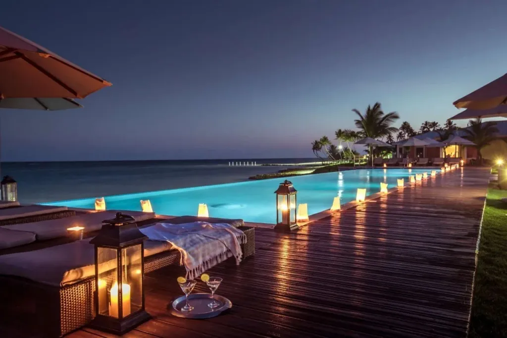Terrasse avec transats et cocktails au bord d'une piscine illuminée en soirée.