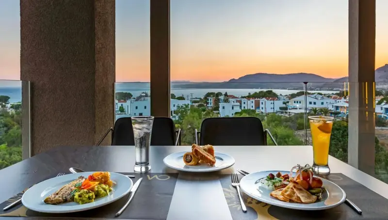 Une table est dressée avec des plats et des boissons face à un paysage côtier au coucher du soleil.