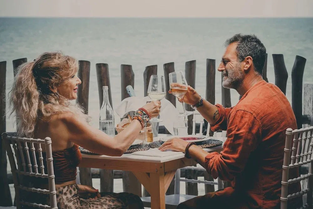 Un couple trinque à un verre de vin assis à une table en extérieur face à la mer.