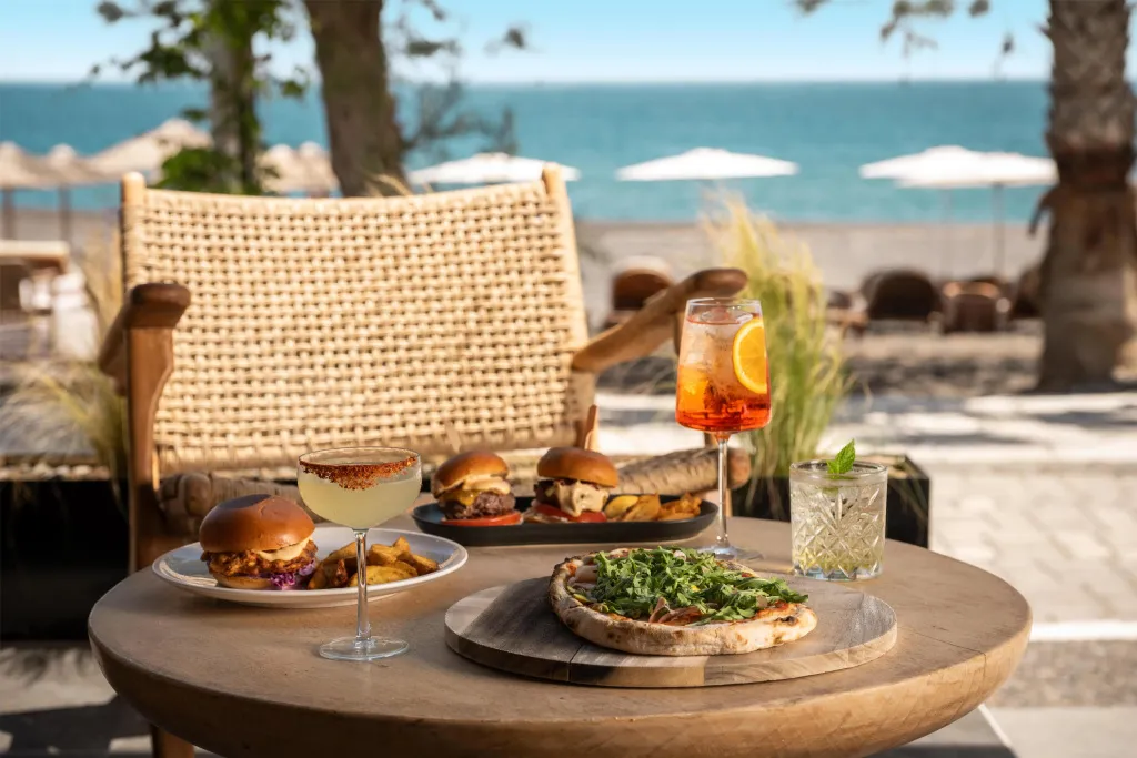 Une table ronde présente des plats et des boissons sur une terrasse ensoleillée avec vue sur la mer.