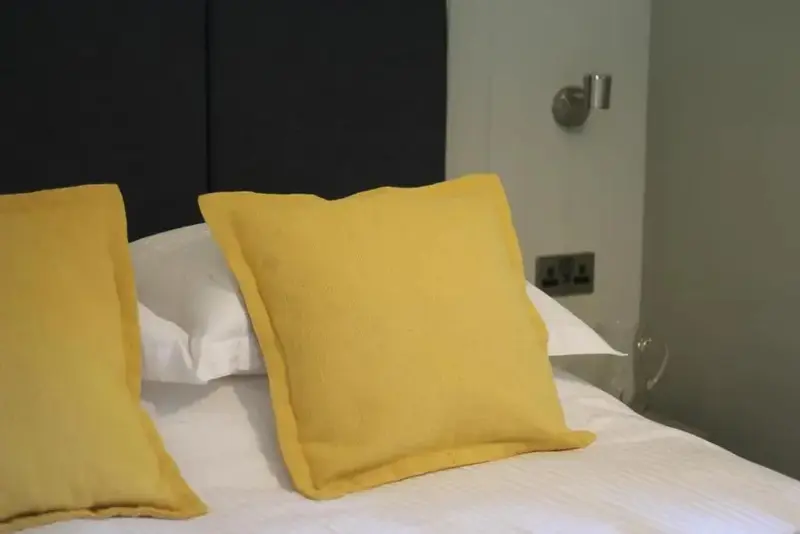 Un lit recouvert de draps blancs et orné de coussins jaunes se trouve contre une tête de lit sombre.