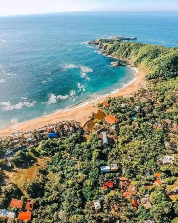 Vue aérienne d'une plage bordée de végétation luxuriante et de bâtiments.