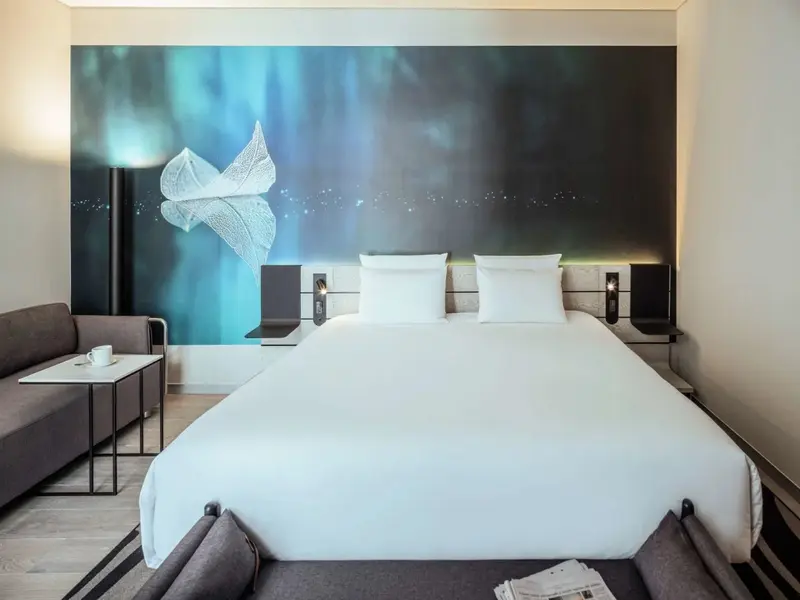 Une chambre lumineuse avec un grand lit et un canapé face à un mur décoré d'une image abstraite.