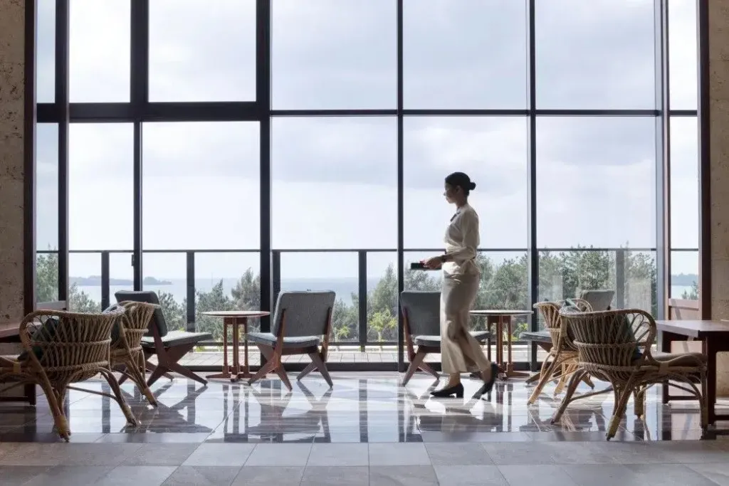 Une femme traverse un espace lumineux avec des fauteuils et une vue sur la mer.