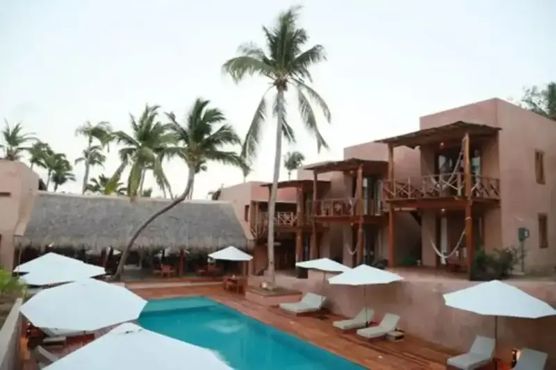Une piscine turquoise bordée de transats blancs et de parasols, devant un bâtiment aux tons chauds.