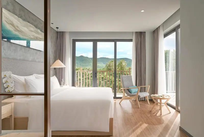 Une chambre lumineuse avec un grand lit et un accès à un balcon offrant une vue sur la végétation.