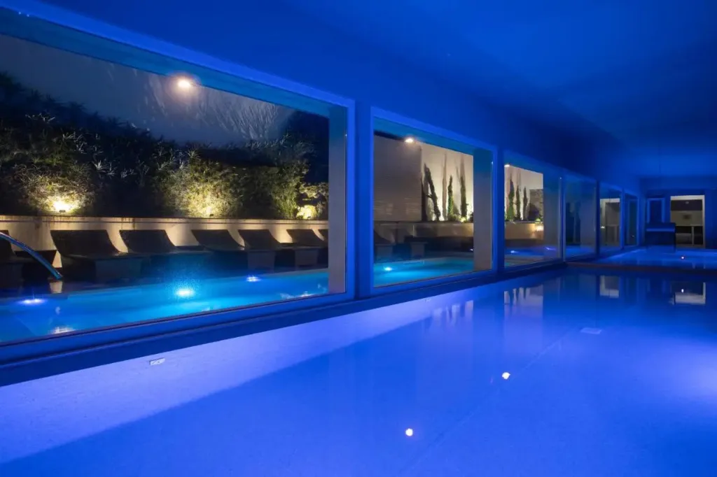Une piscine intérieure illuminée en bleu, avec des transats à l'extérieur.